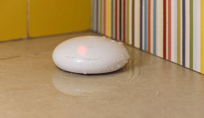 A white oval Fibaro smart flood sensor sitting on a tiled floor with its red indicator light active