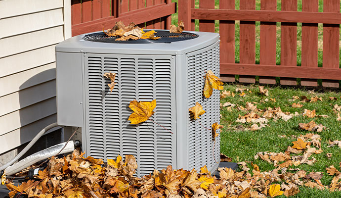 A grey outdoor air conditioning unit surrounded by and covered in dry autumn leaves
