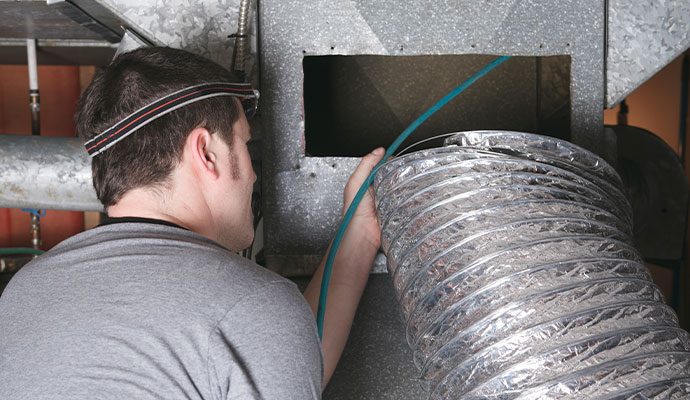 A technician wearing a headlamp inserts a blue fiber-optic camera cable into a silver flexible duct for internal inspection