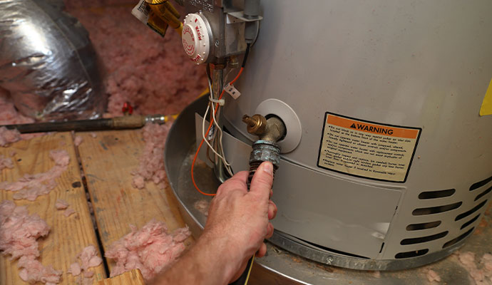 A close-up of a hand attaching a black drainage hose to the brass drain valve at the bottom of a grey water heater