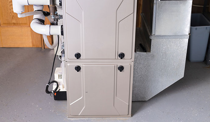 A modern beige indoor furnace unit installed in a garage with white PVC exhaust pipes