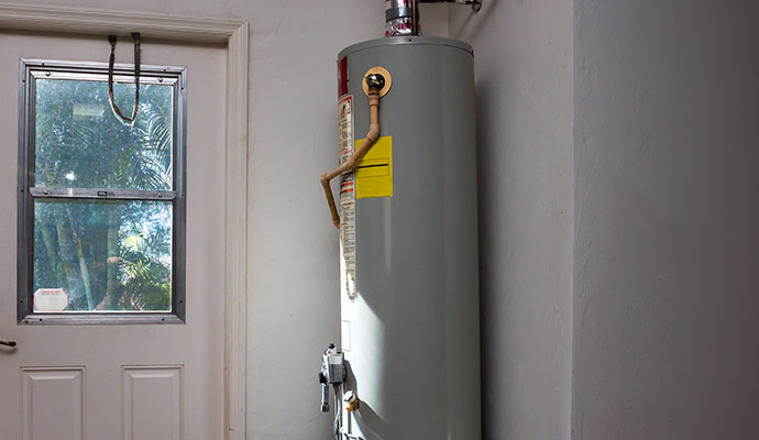 A tall, grey cylindrical gas water heater installed in the corner of a white-walled room