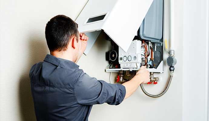 A technician removes the front cover of a white wall-mounted gas boiler to inspect the internal components
